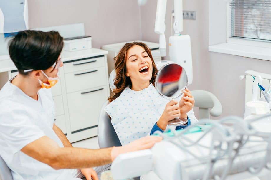 A woman in a dental office inspects her teeth in a hand mirror